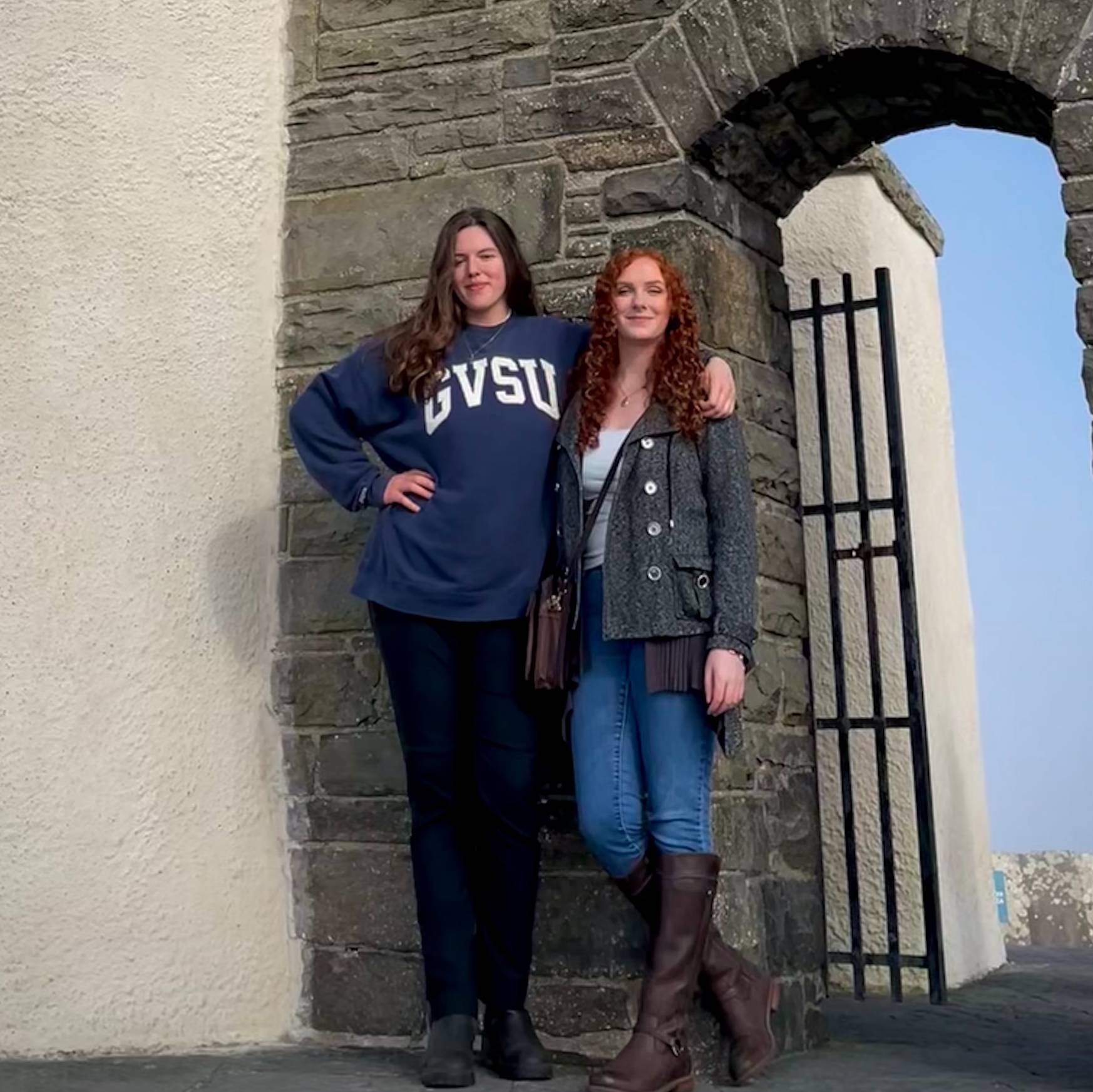 Two alumni pose at an ancient stone wall in Ireland. One wears a GVSU shirt
